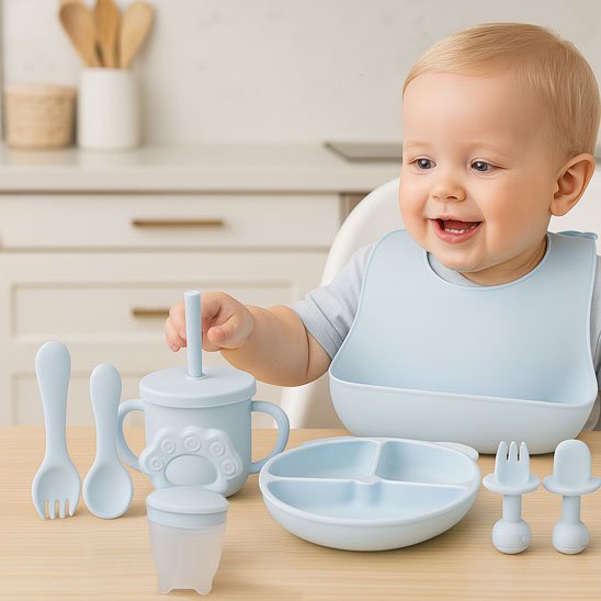 Bébé souriant apprenant à manger seul avec un set de vaisselle en silicone bleu, portant un bavoir et utilisant une tasse à paille et des couverts adaptés
