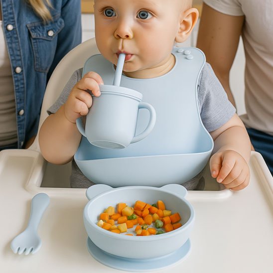 Bébé utilisant une tasse à paille en silicone bleu avec bavoir assorti, bol à ventouse rempli de légumes, apprentissage de l'autonomie au repas